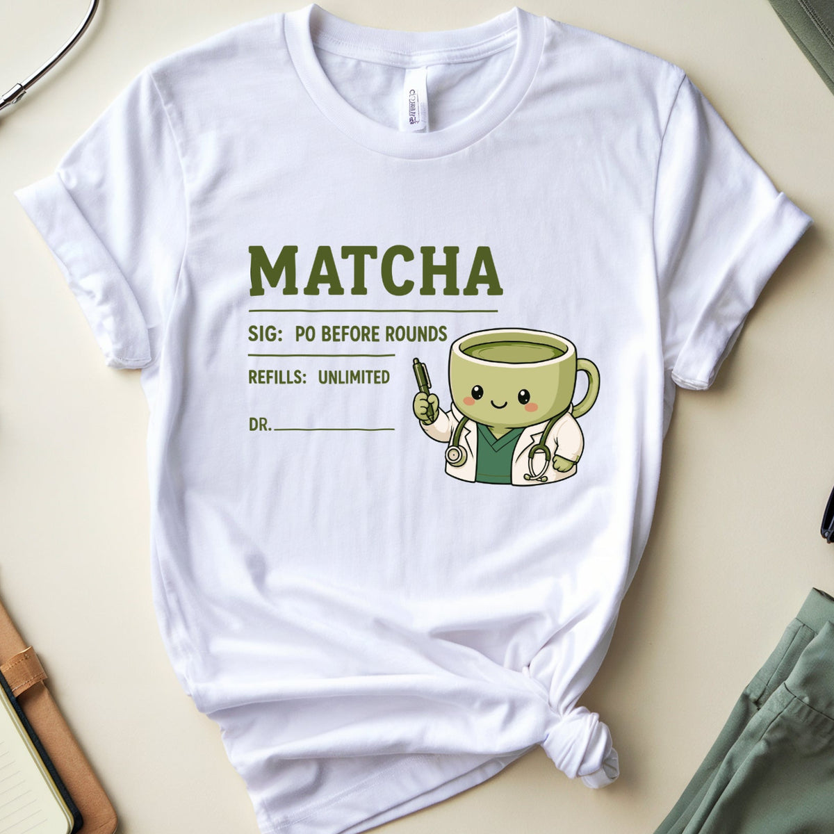 A white graphic t-shirt featuring a cute cartoon matcha tea cup character and the word 'MATCHA' in green, styled in a flat lay with a stethoscope, notebook, glasses, and green pants on a beige background.