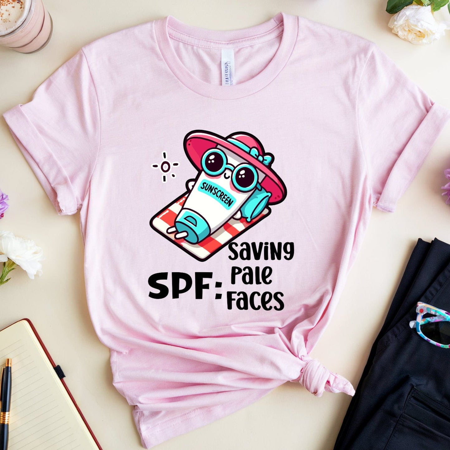 Pink t-shirt with cartoon character and 'Saving Pale Faces' text on a light surface with flowers and a drink.