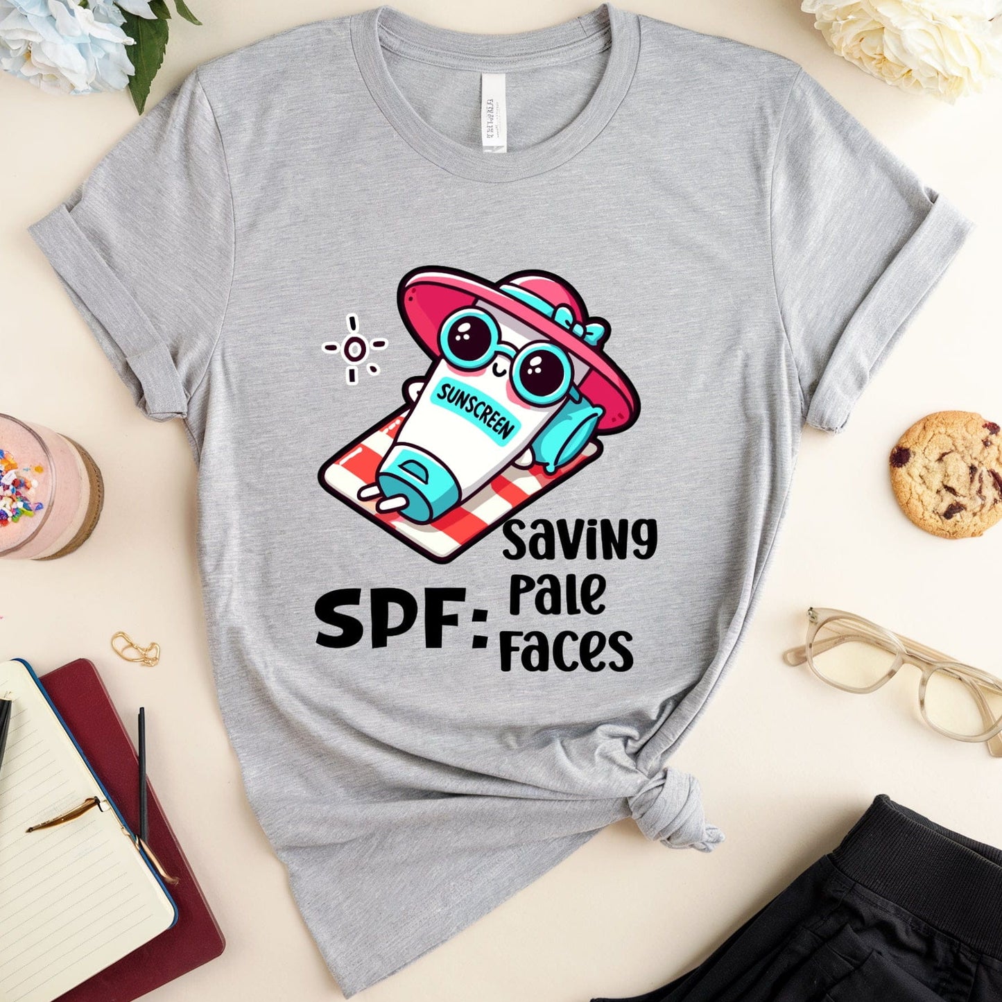 Gray t-shirt with cartoon character and 'SPF: Saving Pale Faces' text on a light surface with accessories.