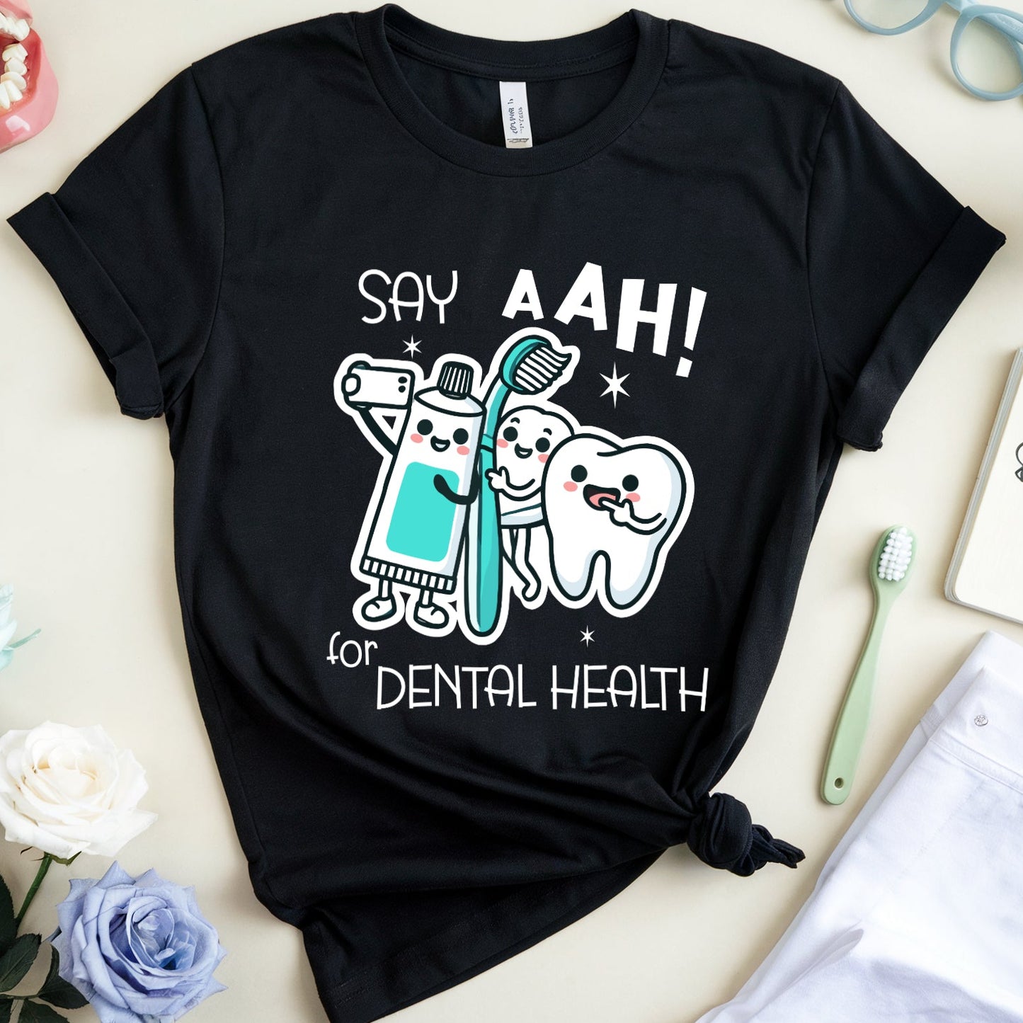A black graphic T-shirt laid flat on a light beige surface, surrounded by dental-themed props including a toothbrush, notebook, glasses, flowers, and a small model mouth. The shirt graphic reads “SAY AAH! for DENTAL HEALTH” with cartoon tooth and brush characters.