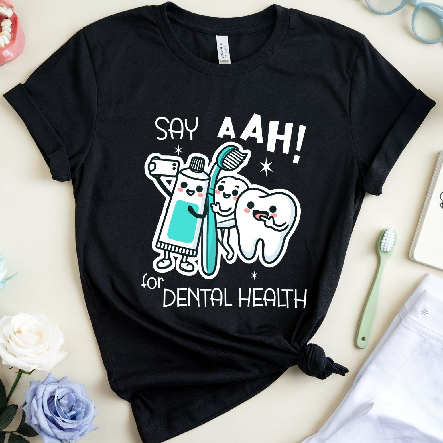 A black graphic T-shirt laid flat on a light beige surface, surrounded by dental-themed props including a toothbrush, notebook, glasses, flowers, and a small model mouth. The shirt graphic reads “SAY AAH! for DENTAL HEALTH” with cartoon tooth and brush characters.