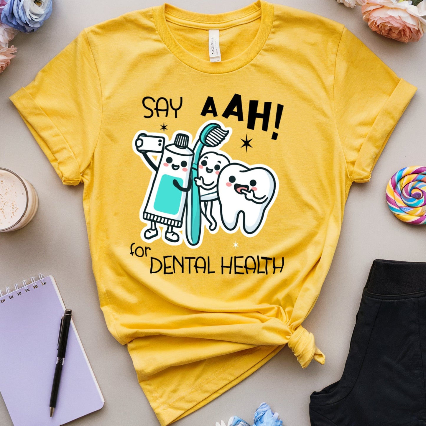 A yellow graphic T-shirt laid flat on a light gray surface, surrounded by flowers, glasses, a notebook, a drink, a lollipop, and black pants. The shirt has a cartoon toothbrush-and-tooth design with the text 'SAY AAH! for DENTAL HEALTH'.