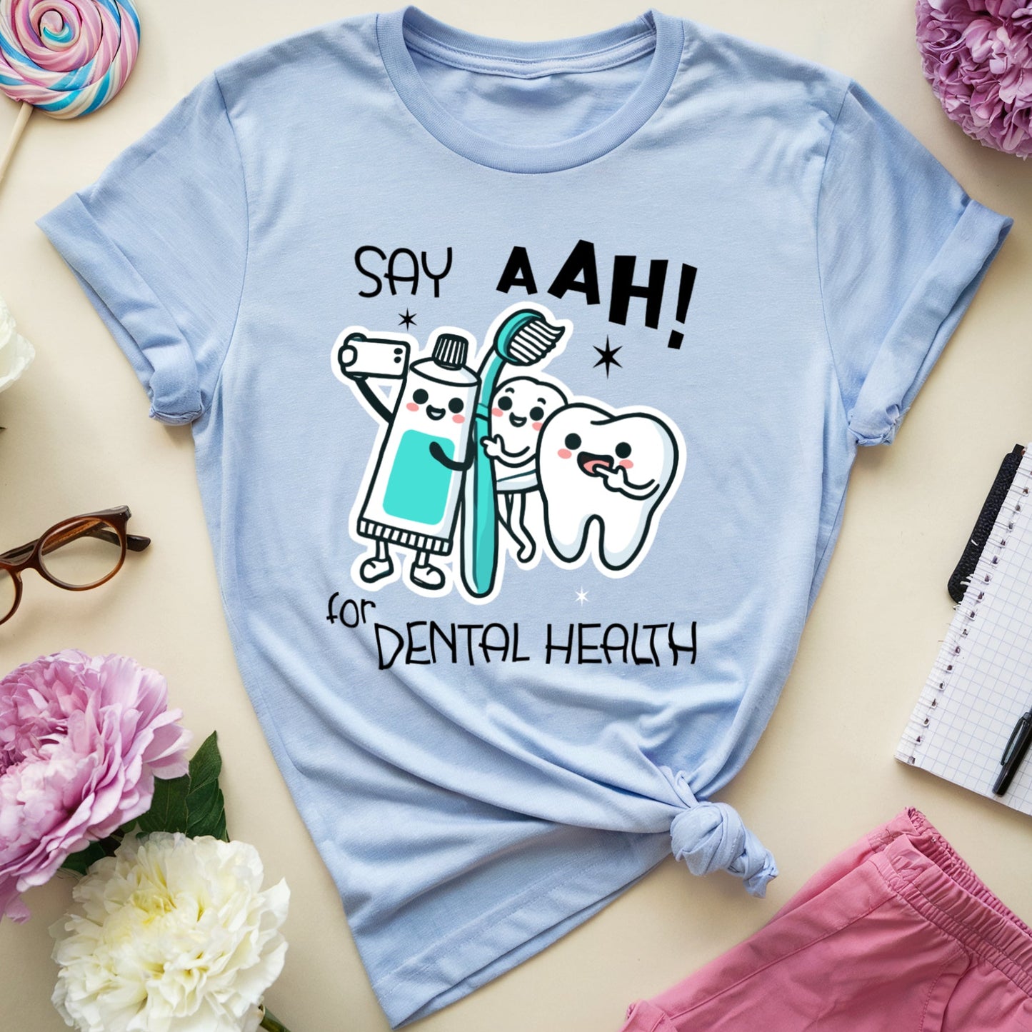 A light blue graphic t-shirt with rolled sleeves, featuring a cartoon illustration of a toothbrush, a tooth, and a dentist holding a sign that says 'SAY AAH! FOR DENTAL HEALTH'. The t-shirt is styled in a flat-lay arrangement with pink and white peonies, eyeglasses, a lollipop, a notebook, and pink shorts on a beige background.