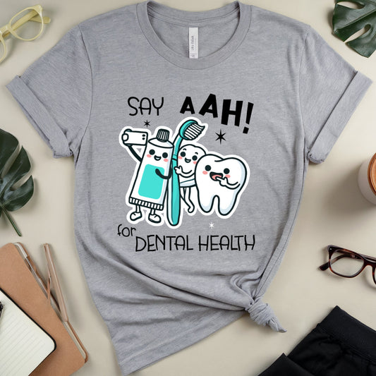 A heather gray graphic t-shirt with a dental-themed cartoon design, styled in a flat-lay arrangement with glasses, a coffee cup, notebook, and leaves on a beige background.