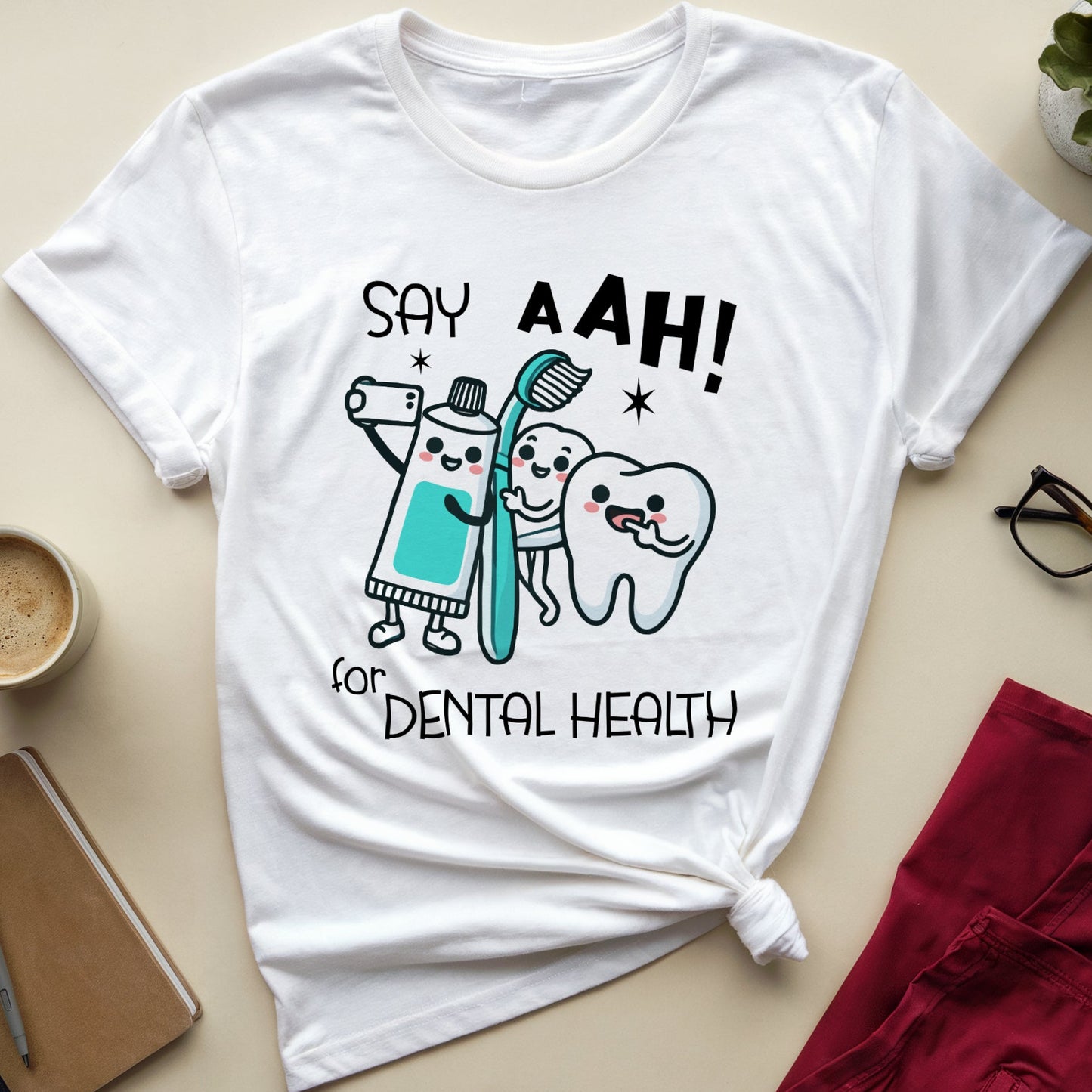 A white graphic t-shirt with a dental health theme, styled in a flat-lay arrangement on a beige surface with a coffee cup, notebook, glasses, a plant, and folded red pants.