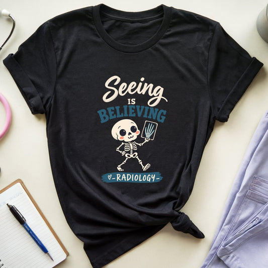 A black graphic t-shirt featuring a cartoon skeleton radiologist with the text 'Seeing IS BELIEVING & RADIOLOGY', styled in a flat lay with medical props including a pink stethoscope, a notebook, a pen, and folded light purple scrubs.