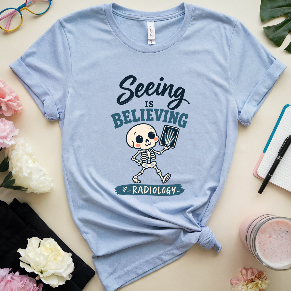 A light blue graphic t-shirt featuring a cartoon skeleton radiologist with the text 'Seeing is BELIEVING & RADIOLOGY'. The t-shirt is styled in a flat-lay arrangement on a beige background with pink and white flowers, a pair of colorful sunglasses, a notebook, a pen, a black cloth, and a glass of pink drink.