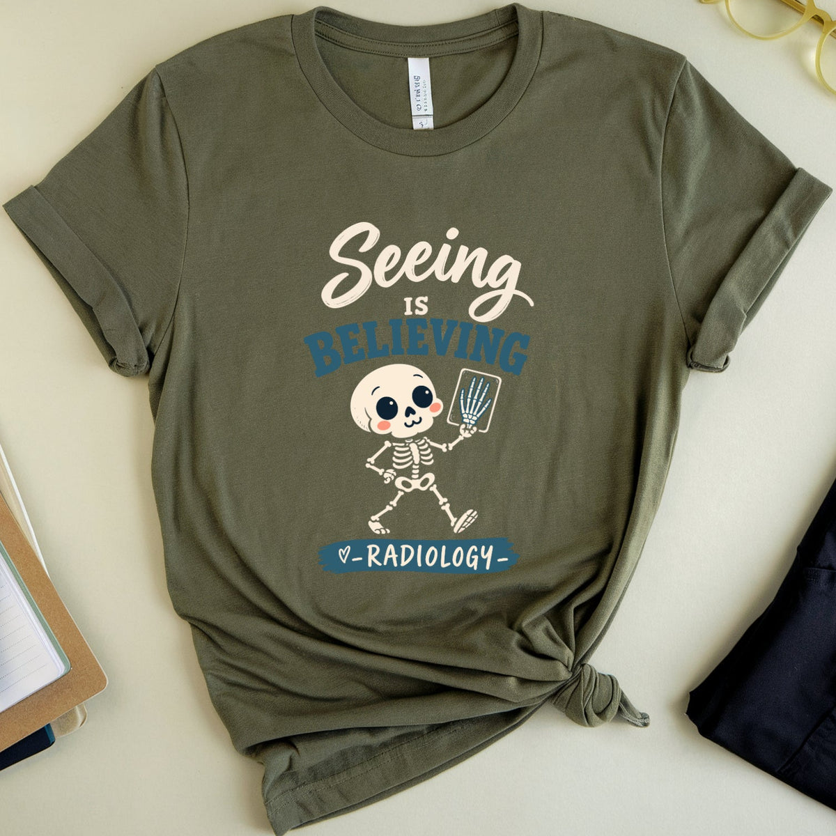 An olive green graphic t-shirt with a rolled hem and front knot, styled in a flat lay with medical scrubs, gold-rimmed glasses, a notebook, and a pen on a light beige background. The t-shirt features a graphic of a skeleton radiologist with the text 'Seeing is BELIEVING' and 'RADIOLOGY'.