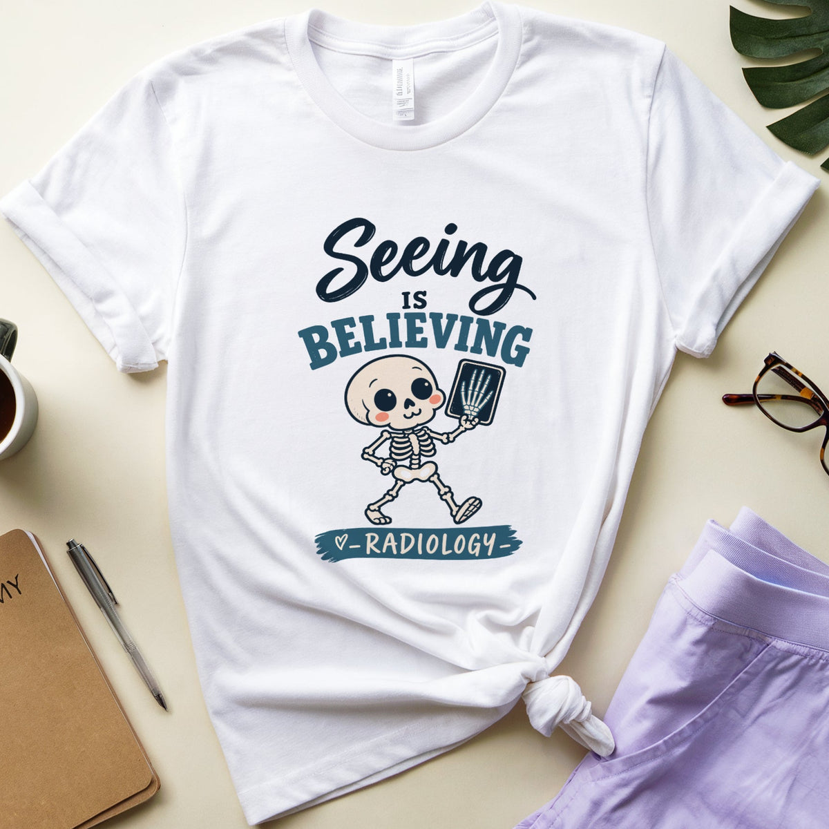 A white graphic T-shirt laid flat on a light beige surface, styled with a cup of coffee, notebook, pen, eyeglasses, and lavender shorts. The shirt has a cartoon skeleton radiologist holding a fluoroscopy tube, with the text 'Seeing is BELIEVING in RADIOLOGY.
