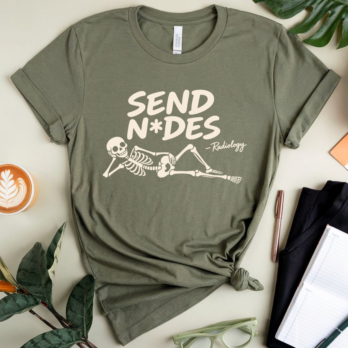 An olive green graphic t-shirt with a skeleton and the text 'SEND N*DES' and 'Rudeology' printed on the front, styled in a flat lay with a coffee cup, green leaves, glasses, a notebook, and pens on a light beige background.