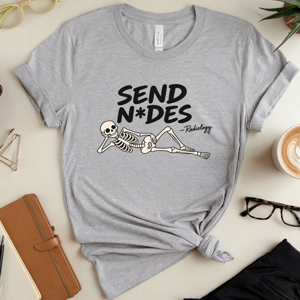 A heather gray graphic T-shirt laid flat on a light beige surface, styled with glasses, a plant, a cup of latte, a notebook, and black pants. The shirt has a black and white graphic of a skeleton lying down with the text 'SEND N*DES -Radiology'.