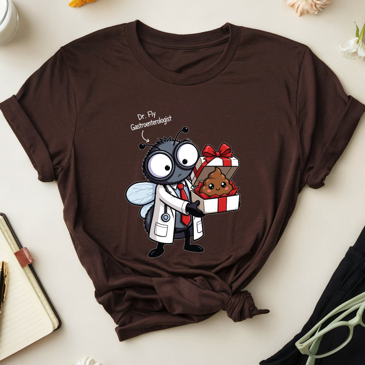 A dark brown graphic t-shirt featuring a cartoon doctor bee holding a poop-shaped gift box, styled in a flat lay with flowers, glasses, a notebook, and a glass of milk on a light beige background.