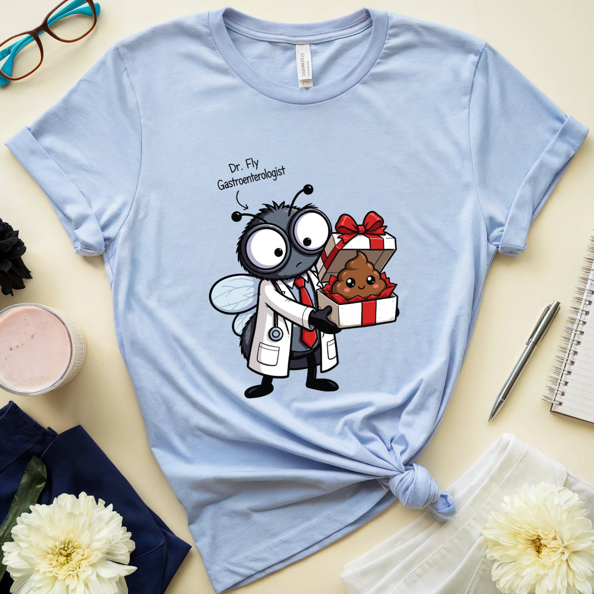 A light blue graphic t-shirt featuring a cartoon bee doctor giving a gift to a poop character, styled in a flat lay with glasses, flowers, a notebook, and a candle on a beige background.