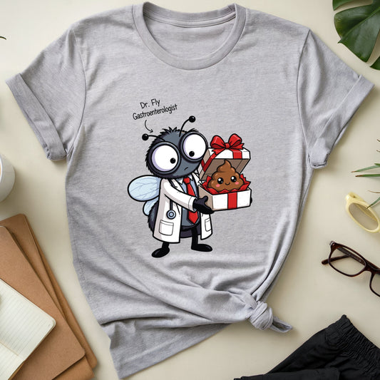 A heather gray short-sleeve T-shirt laid flat on a light beige surface, styled with a coffee cup, notebook, plant leaves, glasses, and folded pants around it. The shirt has a cartoon graphic of a fly dressed as a doctor holding a poop-shaped gift box, with the text 'Dr.