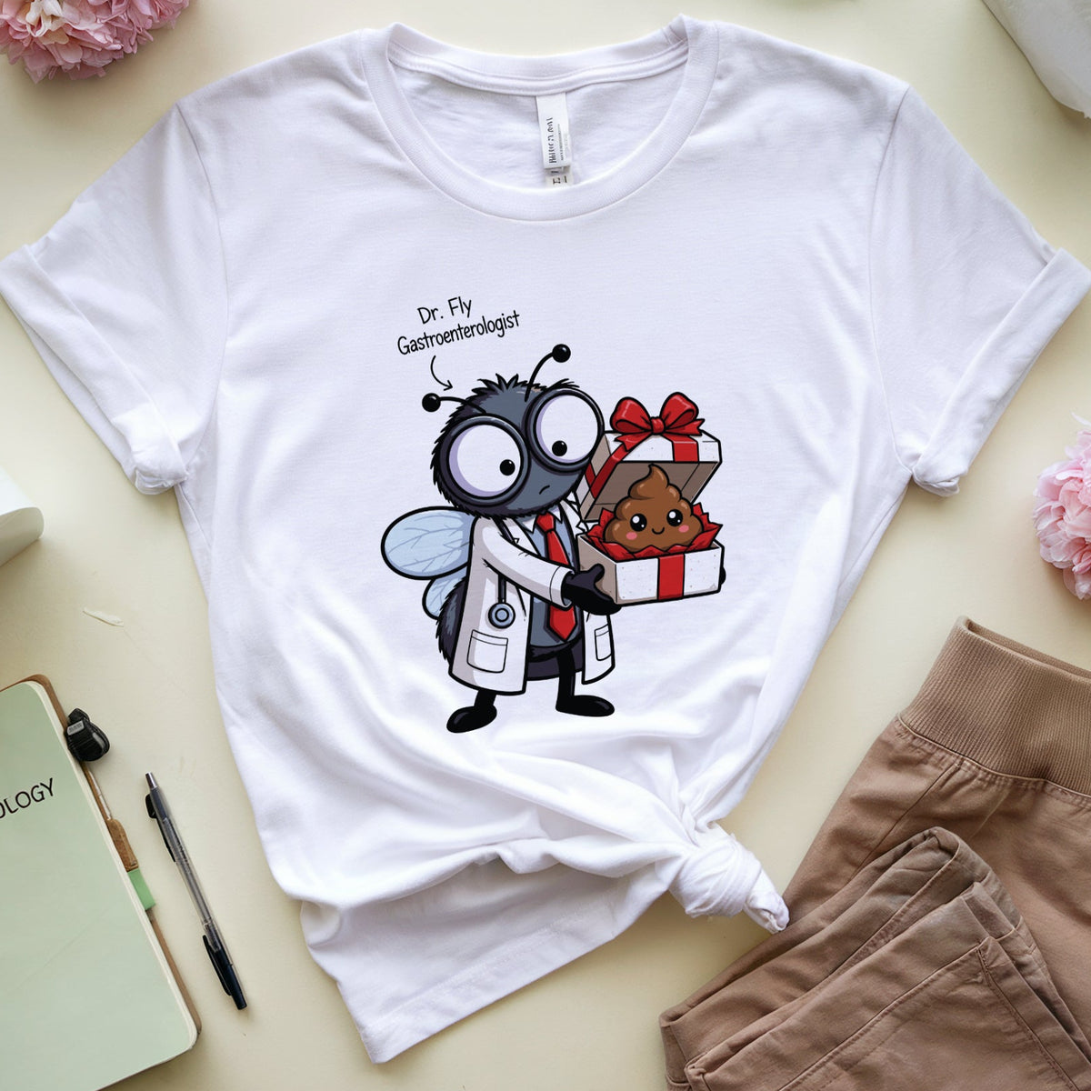 A white graphic t-shirt featuring a cartoon fly dressed as a gastroenterologist holding a gift box, styled in a flat lay with pink flowers, eyeglasses, a tissue, a notebook, and tan shorts on a light beige background.