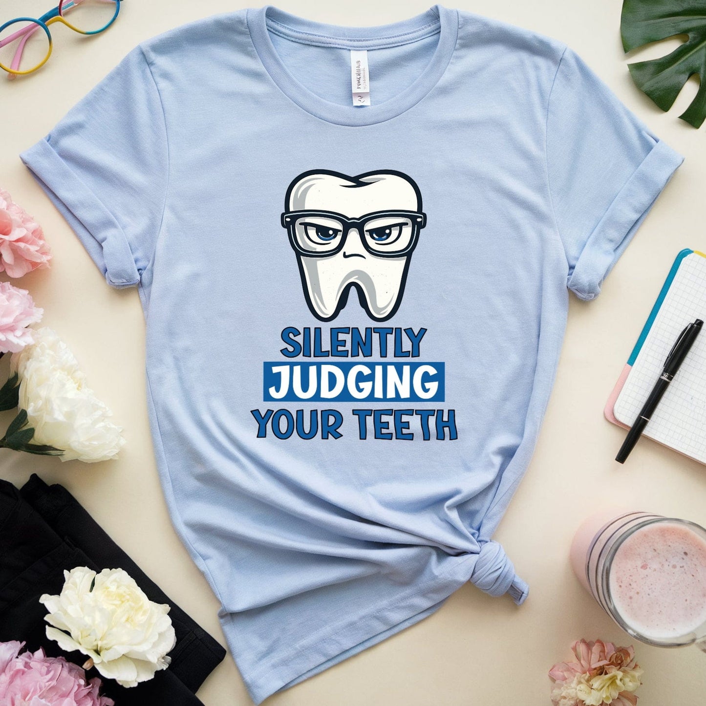 Light blue t-shirt with a graphic of a tooth wearing glasses and text 'Silently Judging Your Teeth' on a table with flowers and a notebook.