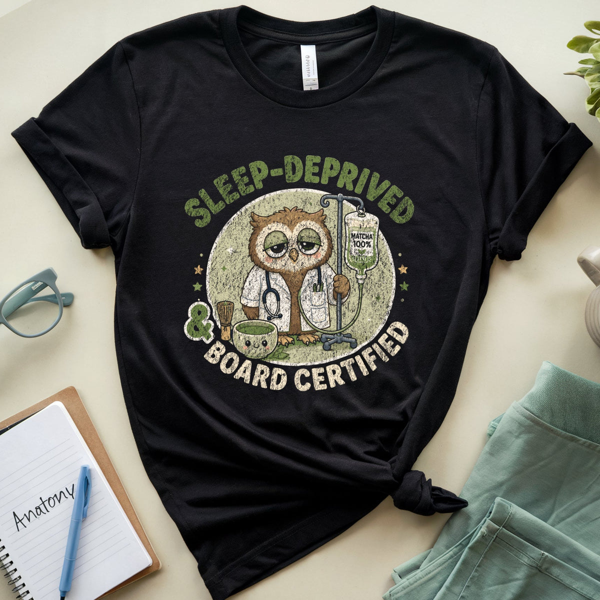 A black graphic t-shirt laid flat on a light beige surface, styled with a notebook, glasses, a mug, and folded mint-green shorts. The t-shirt features a circular owl illustration with a stethoscope and the text 'SLEEP-DEPRIVED BOARD CERTIFIED'.