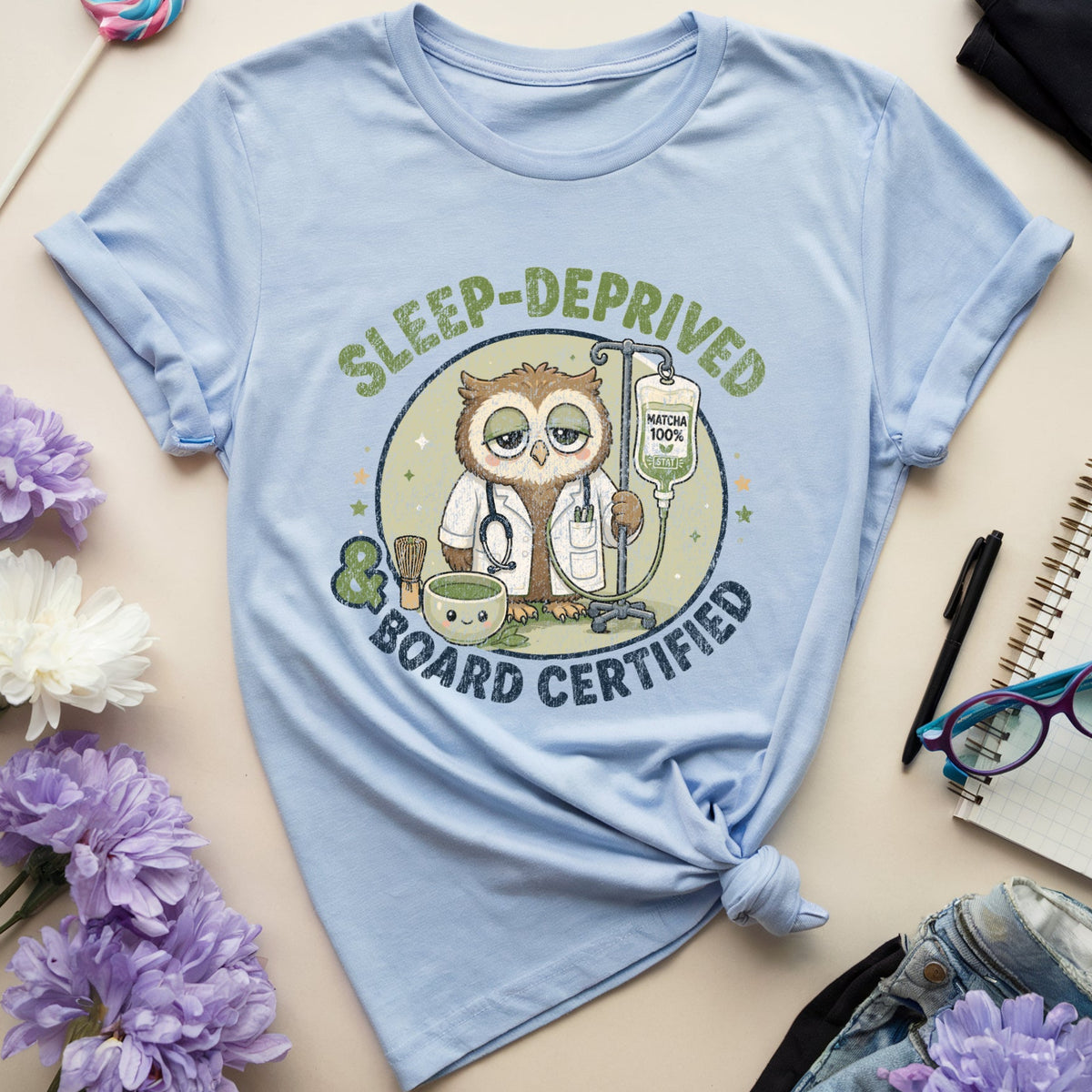 A light blue graphic t-shirt featuring an owl doctor design with the text 'SLEEP-DEPRIVED BOARD CERTIFIED'. The t-shirt is styled in a flat lay with purple and white flowers, a lollipop, eyeglasses, a notebook, and a pen on a beige background.