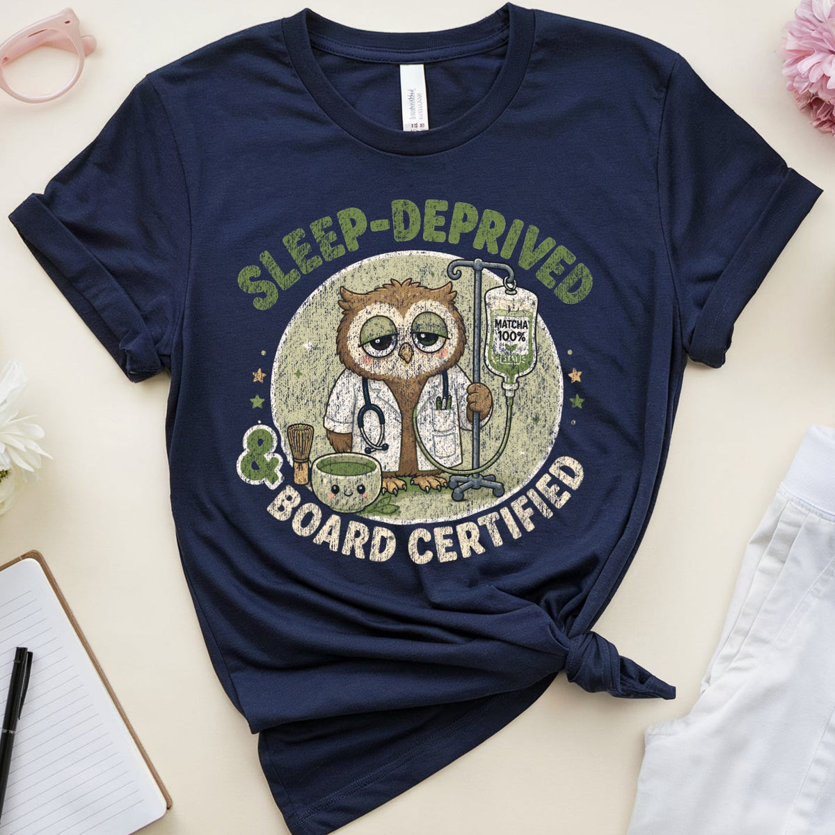A navy blue graphic t-shirt is styled in a flat lay on a light beige background. The shirt features a circular graphic of an owl holding a clipboard with the text 'SLEEP-DEPRIVED BOARD CERTIFIED'.