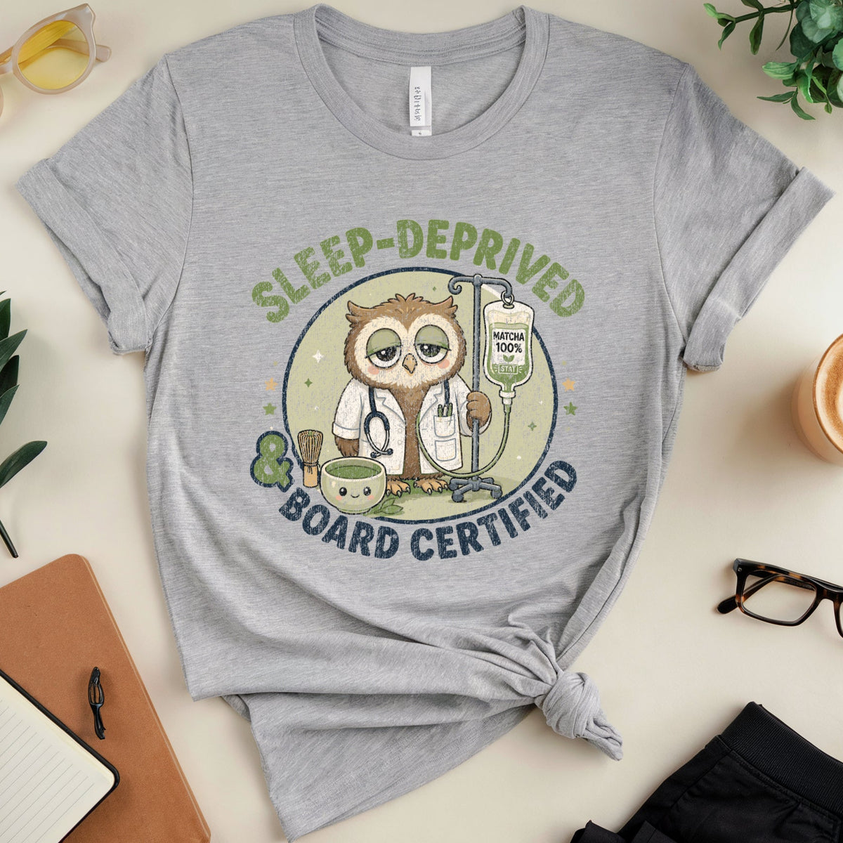 A heather gray graphic t-shirt featuring a cartoon owl dressed as a doctor with the text 'SLEEP-DEPRIVED BOARD CERTIFIED'. The t-shirt is styled in a flat-lay arrangement with a knot at the bottom hem, surrounded by lifestyle props including a notebook, glasses, a coffee cup, and plants on a light beige background.