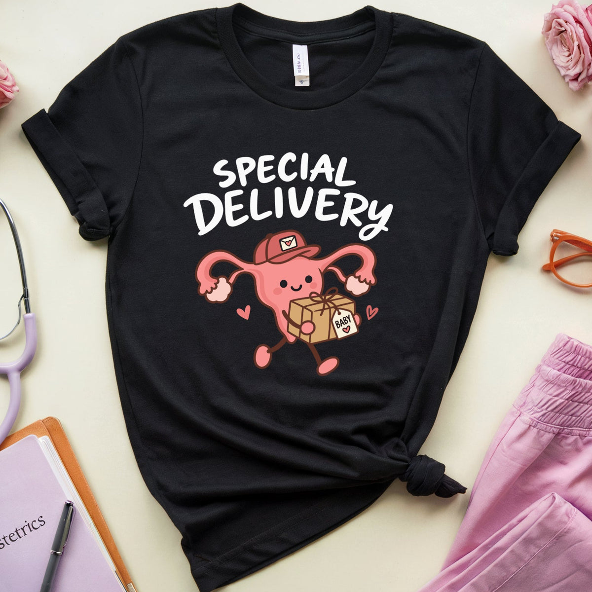 A black graphic t-shirt featuring a pink cartoon obstetrician character with the text 'SPECIAL DELIVERY' in white. The shirt is styled in a flat lay with pink pants, a stethoscope, a notebook, glasses, and roses on a light beige background.