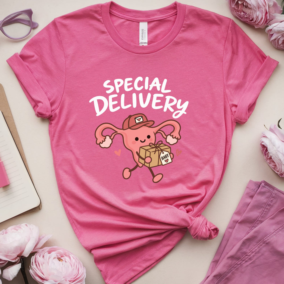 A pink graphic T-shirt laid flat on a light beige surface with pink flowers, glasses, a notebook, and a matching pink garment around it. The shirt has a cartoon uterus delivering a package with the words “SPECIAL DELIVERY” across the front.