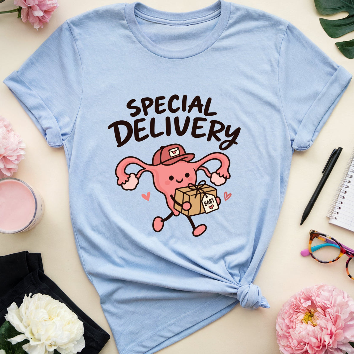 A light blue graphic T-shirt laid flat on a cream surface, styled with flowers, a notebook, glasses, a candle, and black pants. The shirt has a pink cartoon uterus delivering a package with the words “SPECIAL DELIVERY” above it.