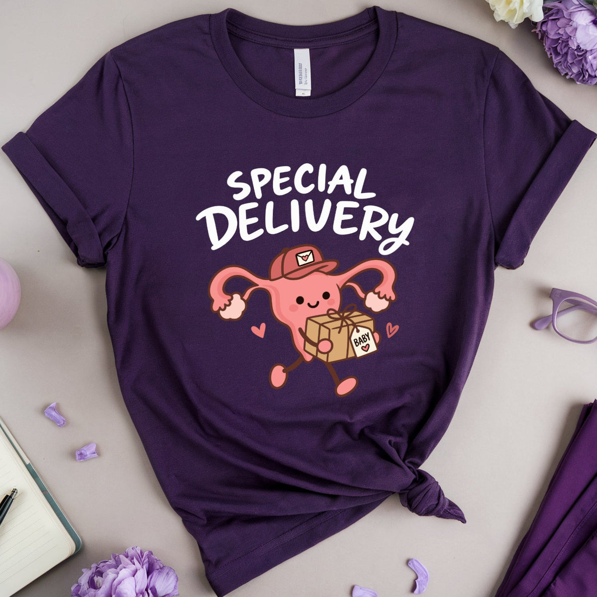 A flat-lay of a dark purple short-sleeve T-shirt with a cartoon graphic of a pregnant uterus delivering a box labeled 'SPECIAL DELIVERY' in white text. The shirt is styled on a neutral beige background with purple and white flowers, glasses, a notebook, and matching purple pants.