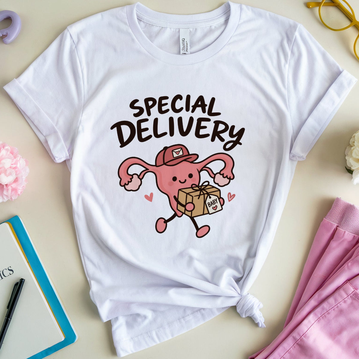A white graphic t-shirt featuring a cartoon uterus holding a baby bag with the text 'SPECIAL DELIVERY' in black. The shirt is styled in a flat lay with pink pants, a purple animal head decor, a pink carnation, a notebook, and yellow glasses on a beige background.