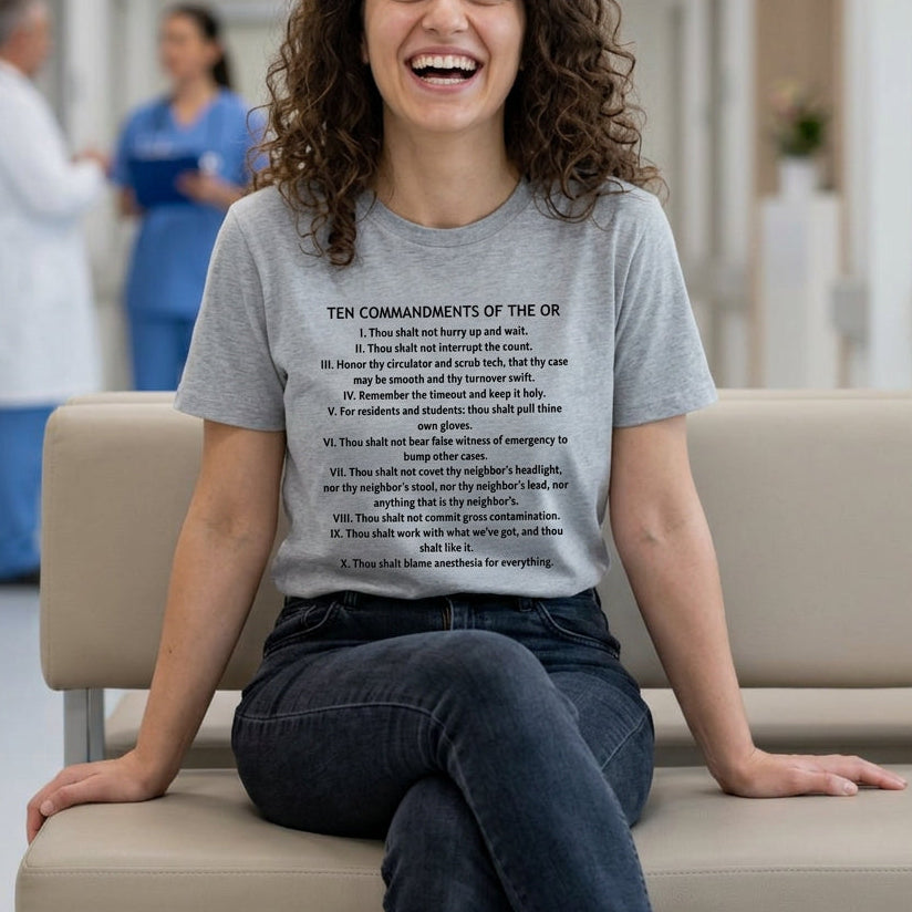 A woman with curly brown hair sits smiling on a beige chair in a brightly lit hospital hallway, wearing a light gray graphic T-shirt with black printed text and dark jeans.