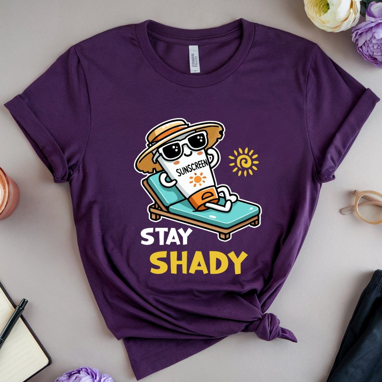 Purple t-shirt with 'Stay Shady' graphic on a table with flowers and a coffee cup.