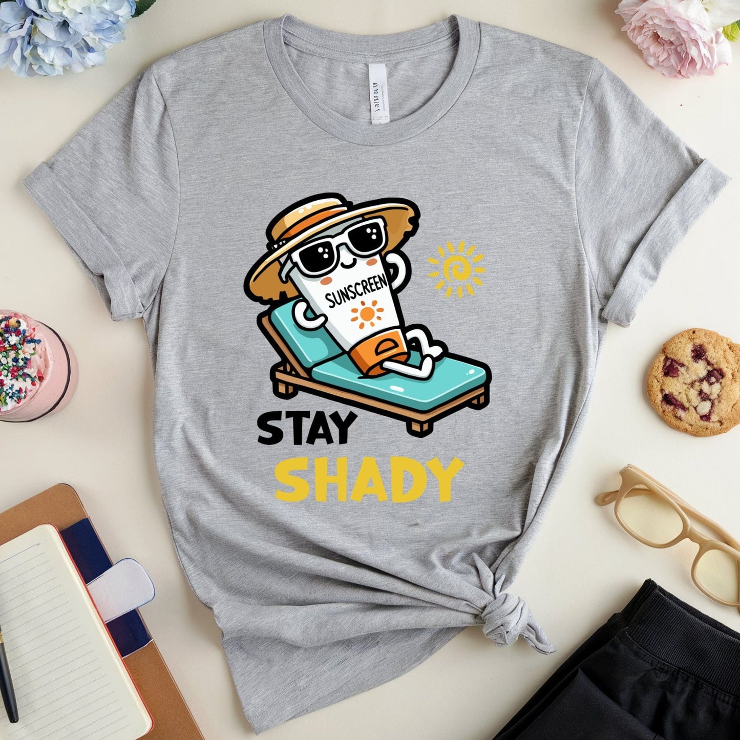 Gray t-shirt with cartoon sun and 'Stay Shady' text on a light surface with accessories.