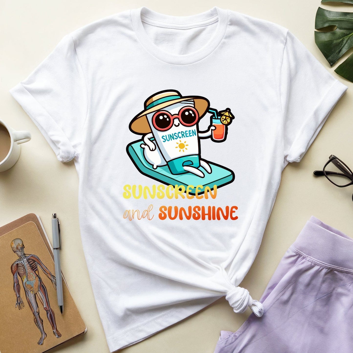 White t-shirt with a cartoon character and 'Sunscreen and Sunshine' text on a light surface with a coffee cup, glasses, and notebook.