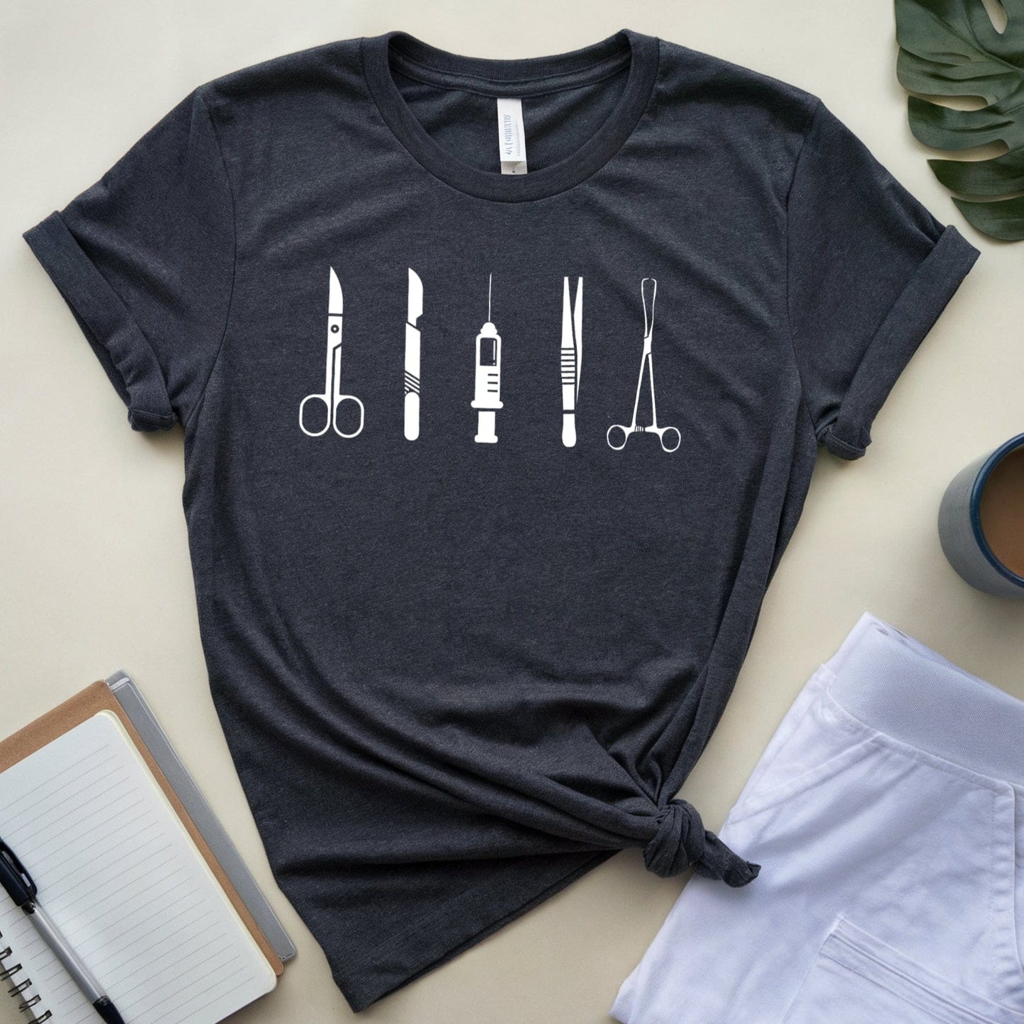 Dark gray t-shirt with medical tool design on a beige surface with notebook, pen, and coffee cup.