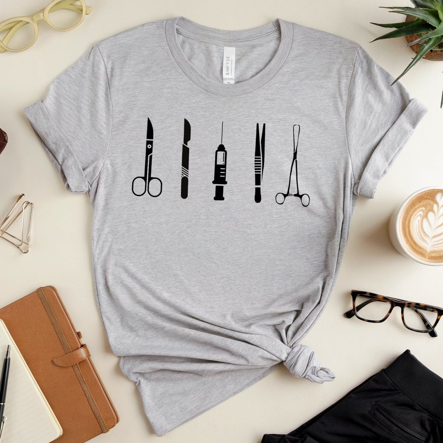 Gray t-shirt with black medical tool icons on a desk with office supplies.