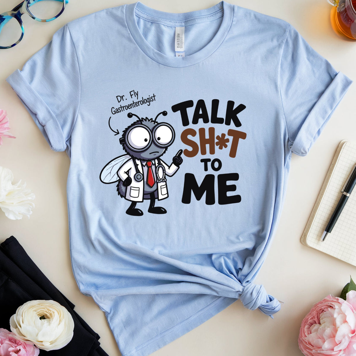 A light blue graphic T-shirt laid flat on a light beige surface, styled with flowers, glasses, a notebook, and a glass of tea. The shirt has a cartoon fly dressed as a doctor with the text 'Dr.
