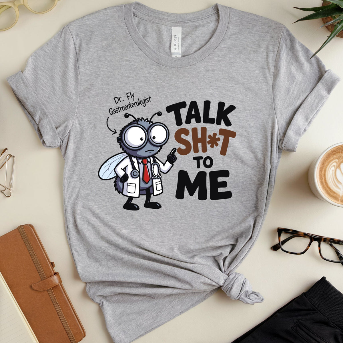 A heather gray graphic T-shirt laid flat on a light beige surface, styled with glasses, a notebook, a cup of latte, and a plant. The shirt features a cartoon fly dressed as a doctor pointing upward beside the text 'Dr.