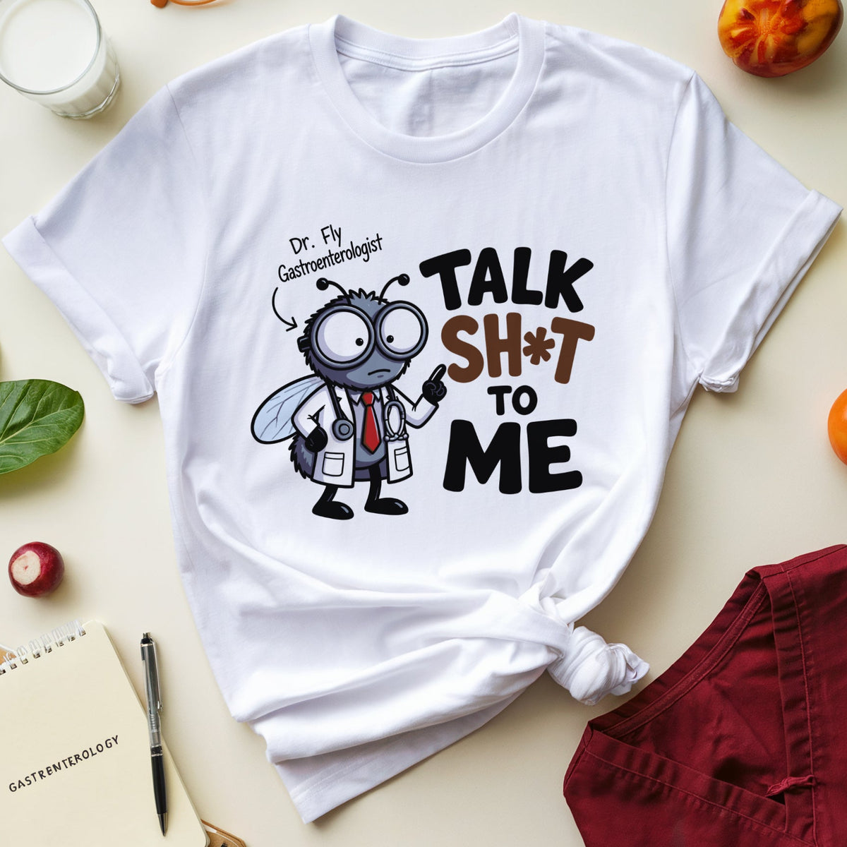 A white graphic T-shirt laid flat on a light beige surface, surrounded by styled props including orange glasses, a glass of milk, green leaves, a notebook, a pen, a radish, and fruit. The shirt has a cartoon fly dressed as a gastroenterologist with the text 'Dr.