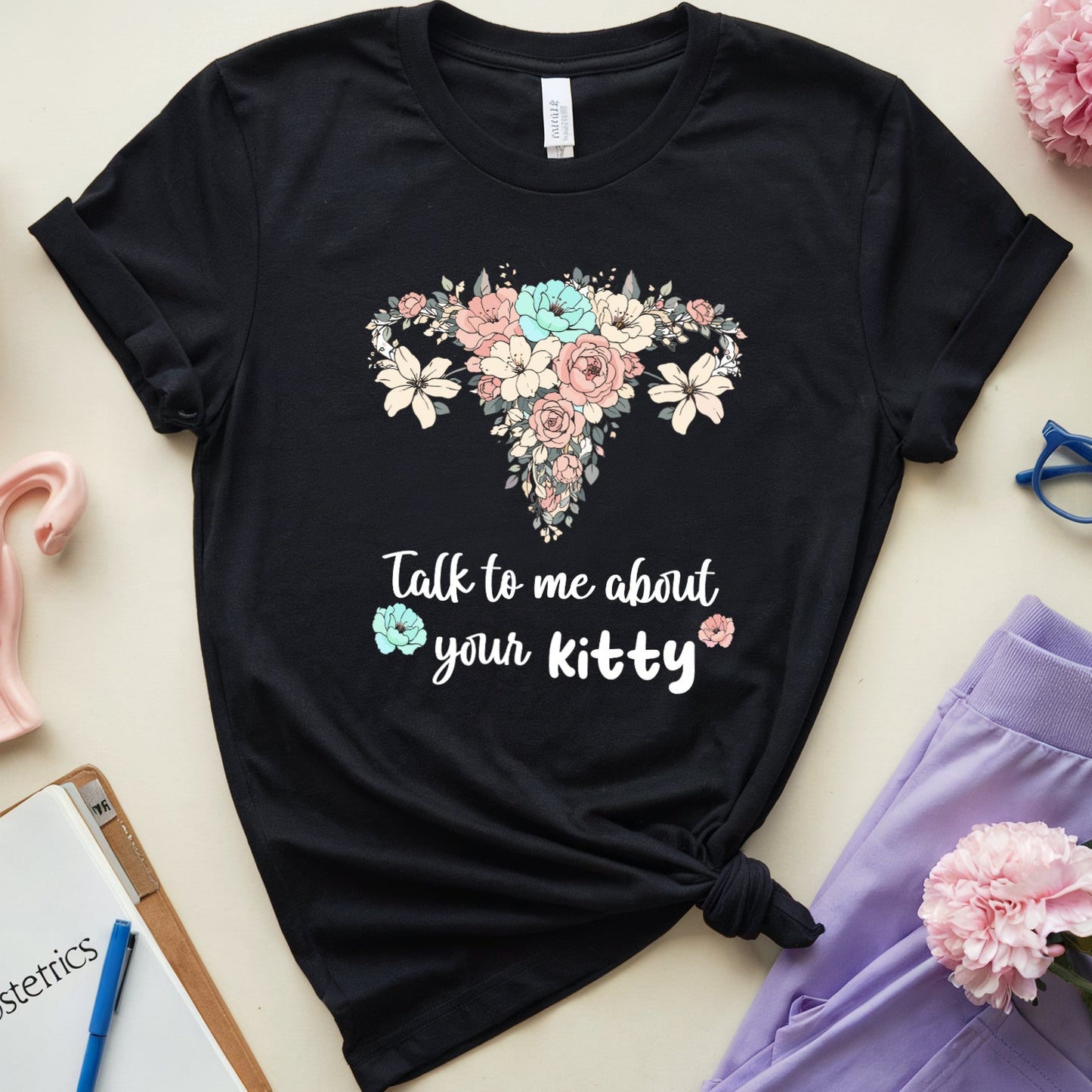Black t-shirt with floral uterus design and text, surrounded by flowers and medical-themed items on a light background.