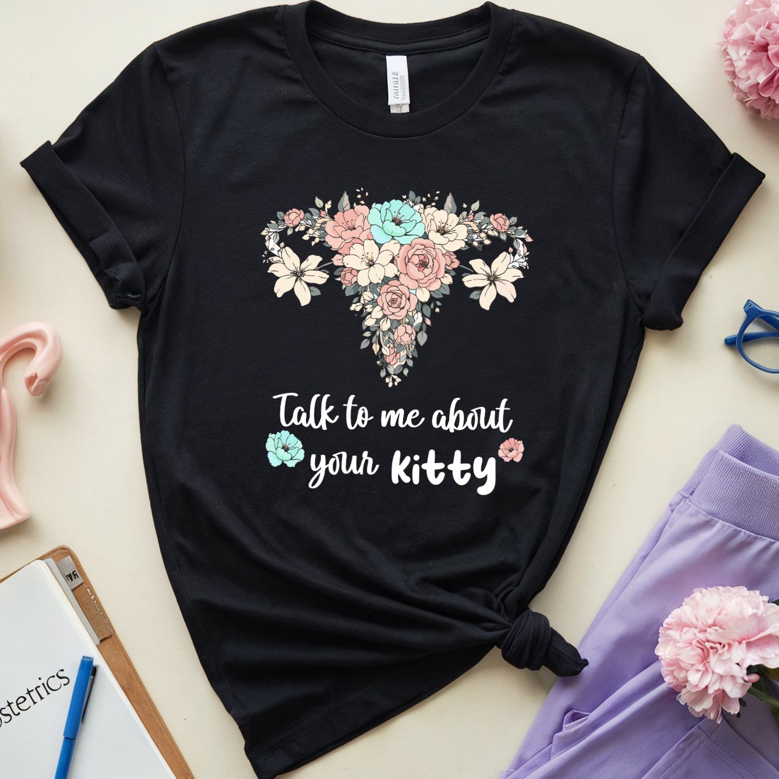 Black t-shirt with floral uterus design and text, surrounded by flowers and medical-themed items on a light background.