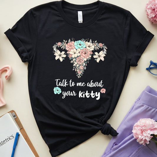 Black t-shirt with floral uterus design and text, surrounded by flowers and medical-themed items on a light background.