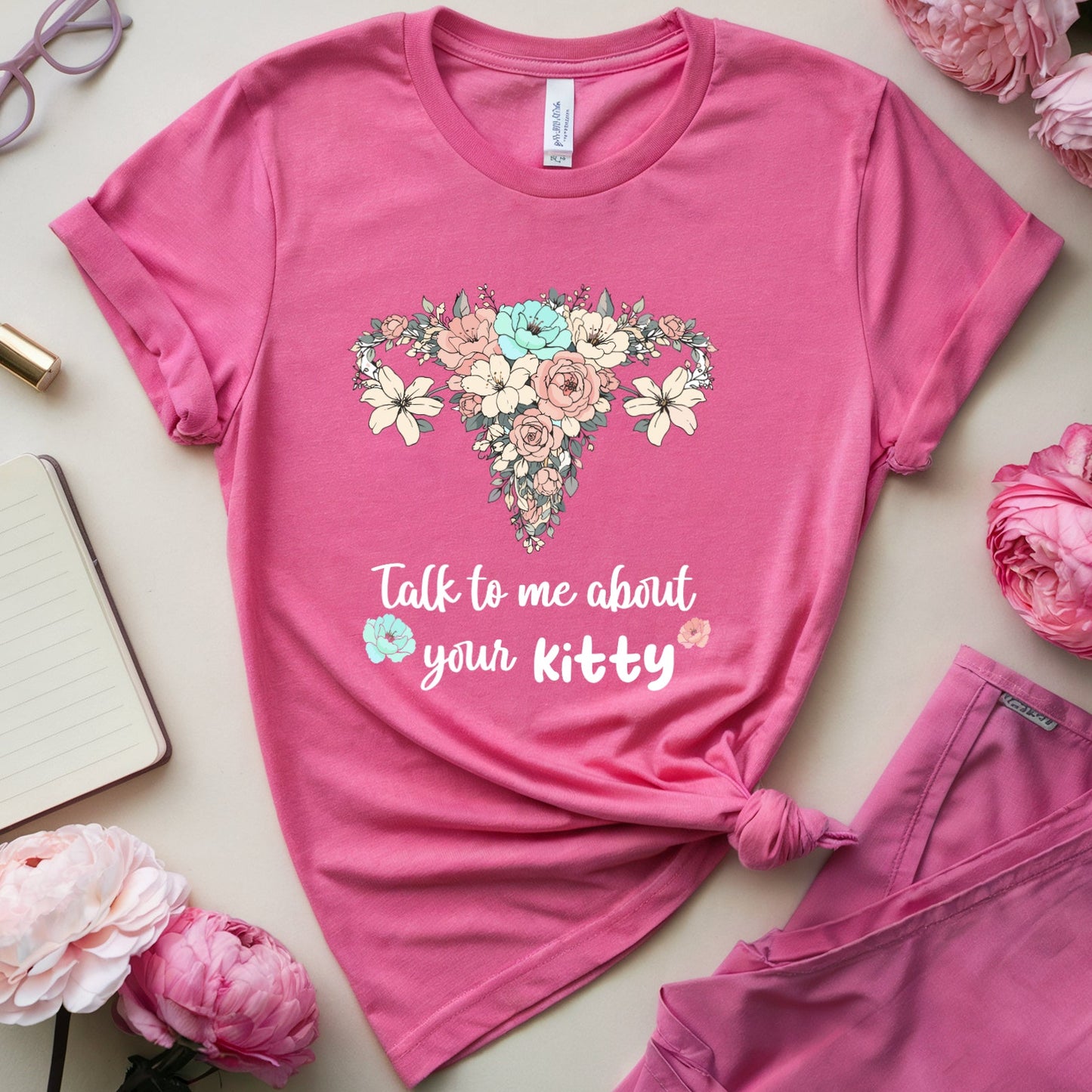 Pink t-shirt with floral design and text, surrounded by pink flowers and accessories on a light surface.