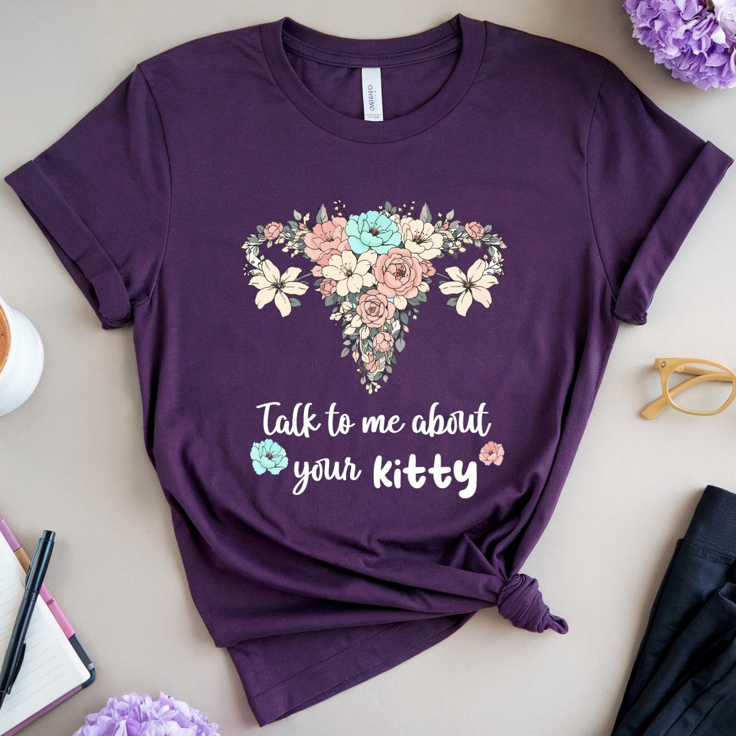 Purple t-shirt with floral design and text on a table with coffee, flowers, and glasses.
