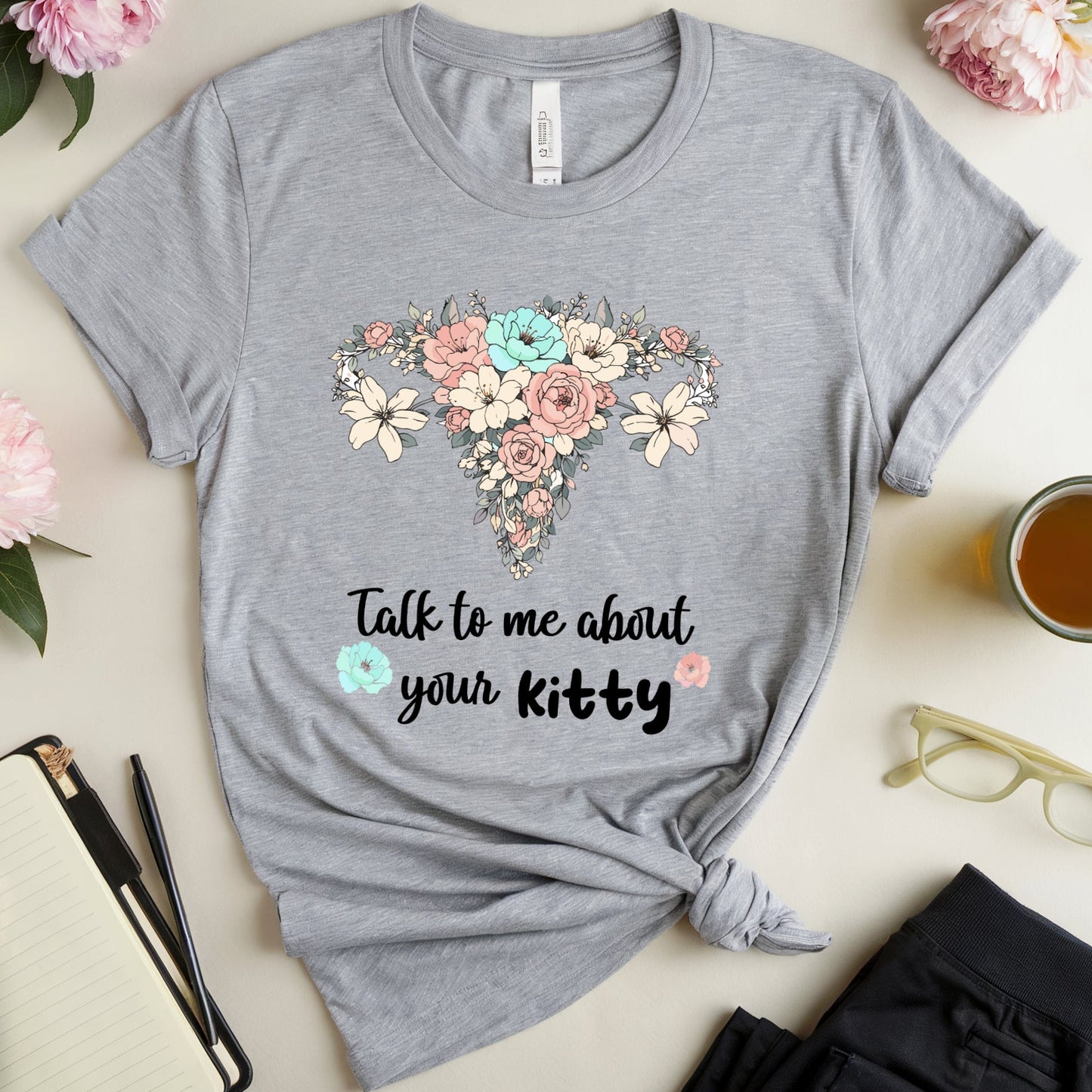 Gray t-shirt with floral design and text on a table with flowers and a cup of tea.