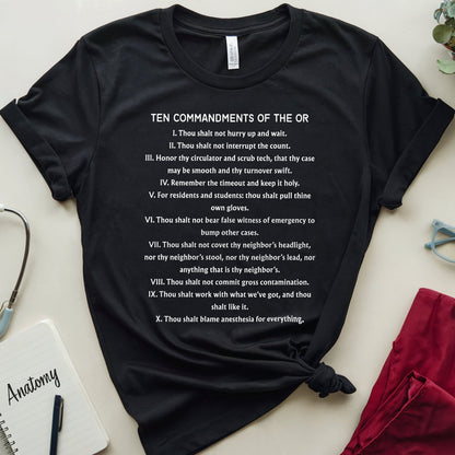 A black graphic t-shirt with white text reading 'TEN COMMANDMENTS OF THE OR' is laid flat on a light beige surface, styled with medical-themed props including a stethoscope, an 'Anatomy' notebook, and a red gynecological exam pad.