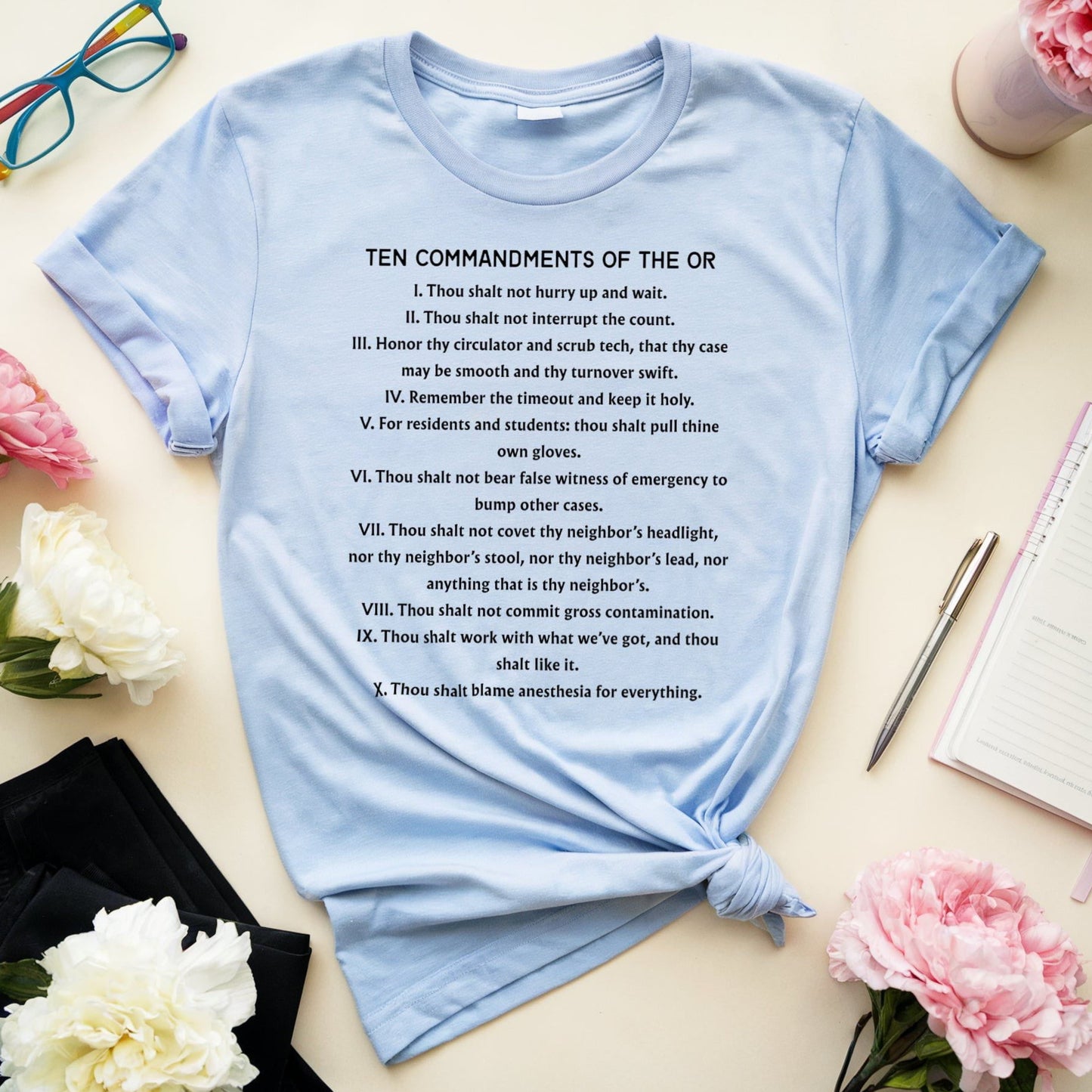 Light blue t-shirt with 'Ten Commandments of the OR' text on a table with flowers and glasses.