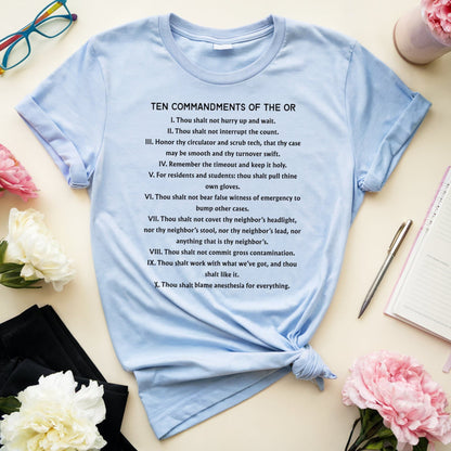 Light blue t-shirt with 'Ten Commandments of the OR' text on a table with flowers and glasses.
