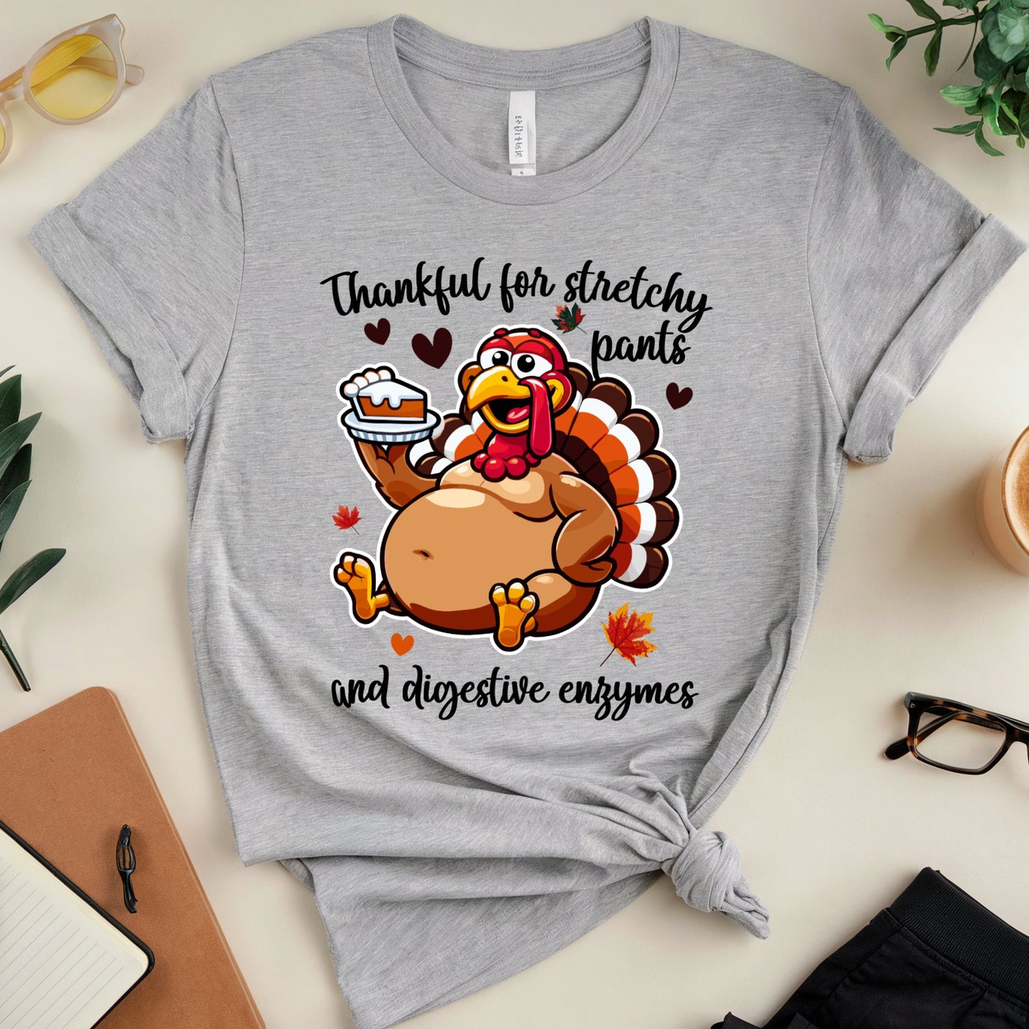 A gray graphic T-shirt featuring a turkey holding pie for Thanksgiving dinner with the words "thankful for stretchy pants ad digestive enzymes"