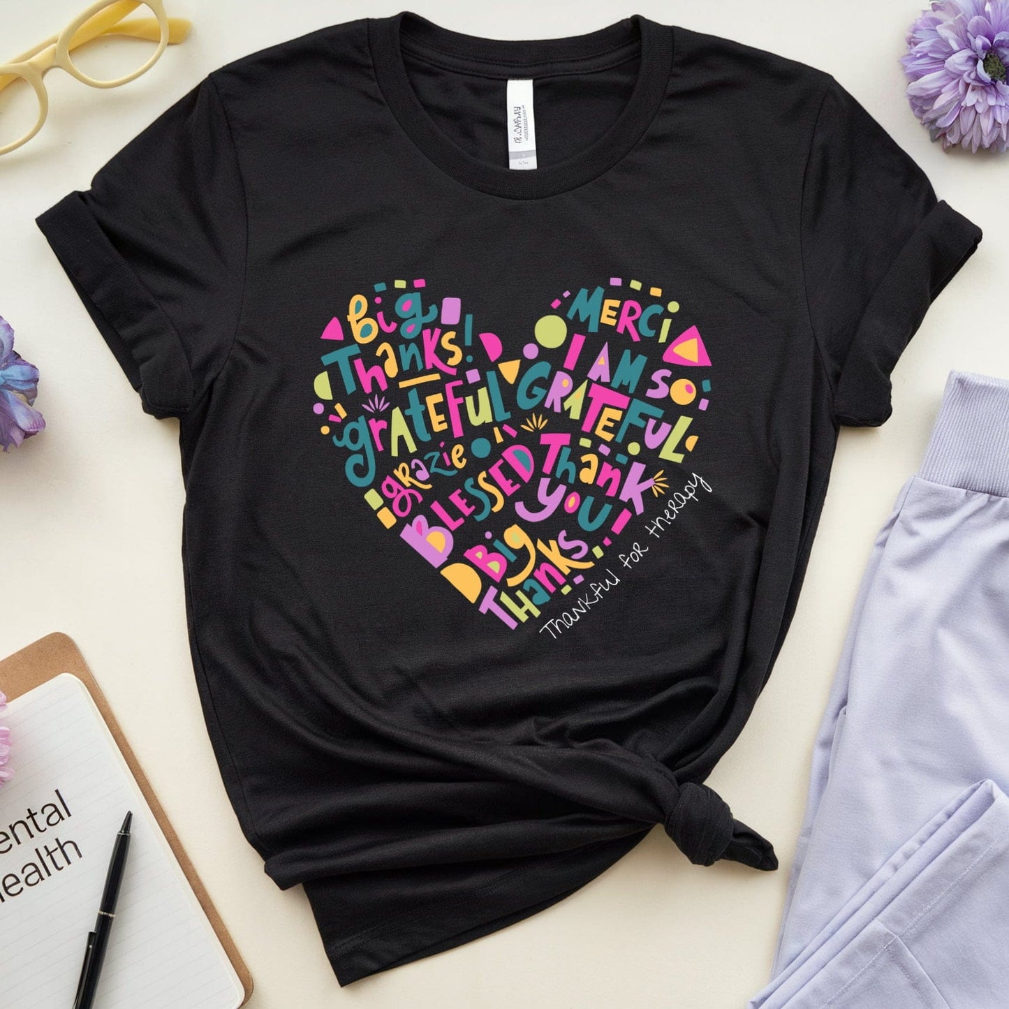 Black t-shirt with colorful heart-shaped text design on a light surface with flowers and glasses.