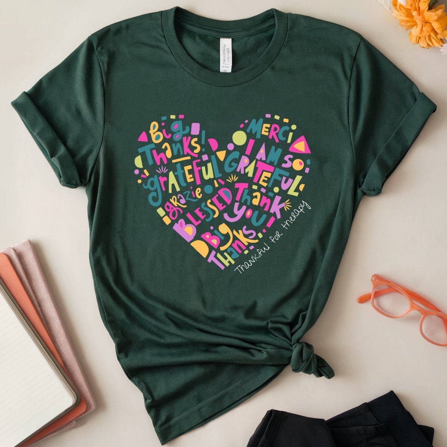 Green t-shirt with colorful heart-shaped text design on a light surface with flowers and glasses.